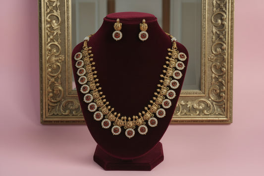 Gold necklace with red stones and matching earrings on a white stand against a pink background