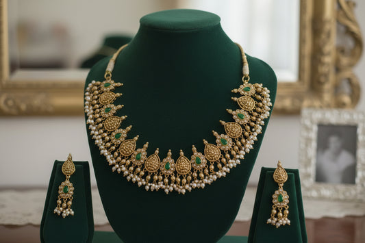 Gold necklace with green stones and matching earrings on a green stand against a light purple background