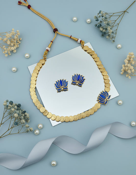 Gold necklace with blue floral designs on a white card, surrounded by pearls and dried flowers on a light blue background.