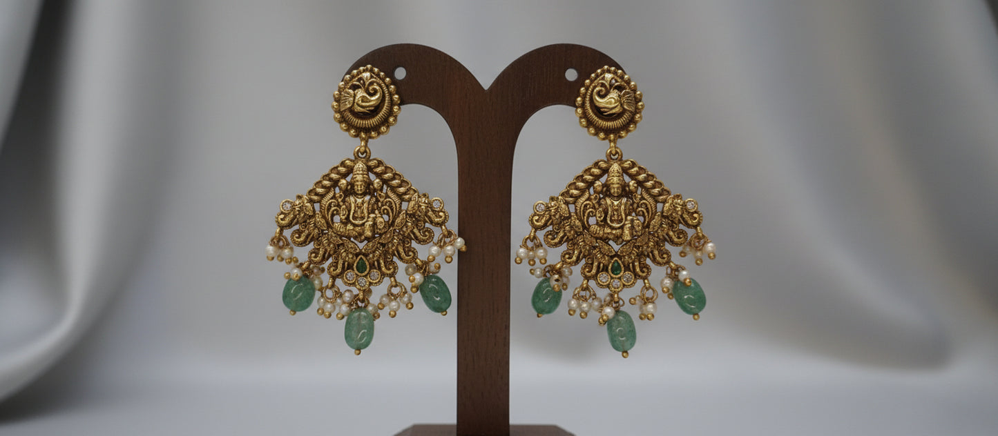 Gold earrings with green stones on a brown stand against a gray background