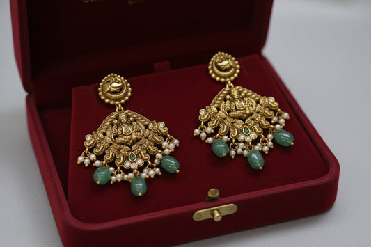 Gold earrings with green stones on a brown stand against a gray background