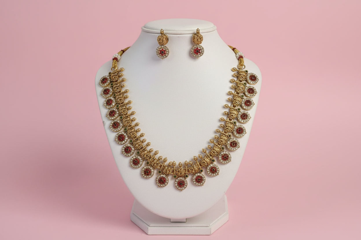 Gold necklace with red stones and matching earrings on a white stand against a pink background