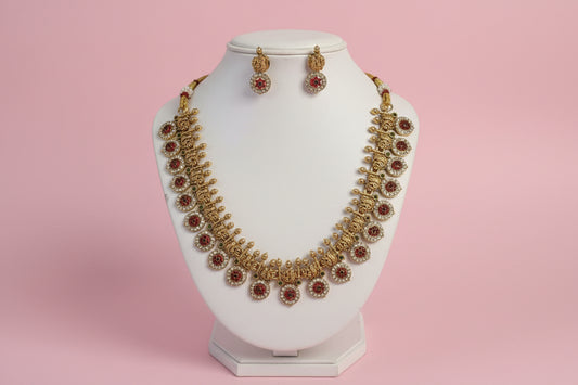 Gold necklace with red stones and matching earrings on a white stand against a pink background