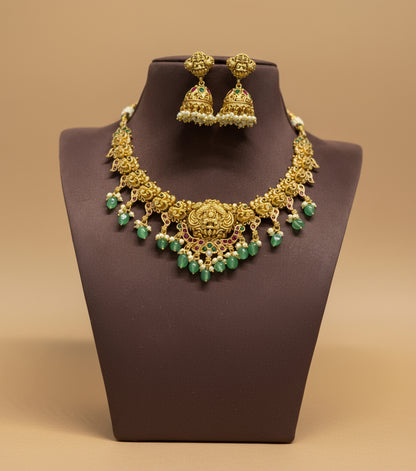 Gold necklace with green beads and matching earrings on a brown stand against a beige background
