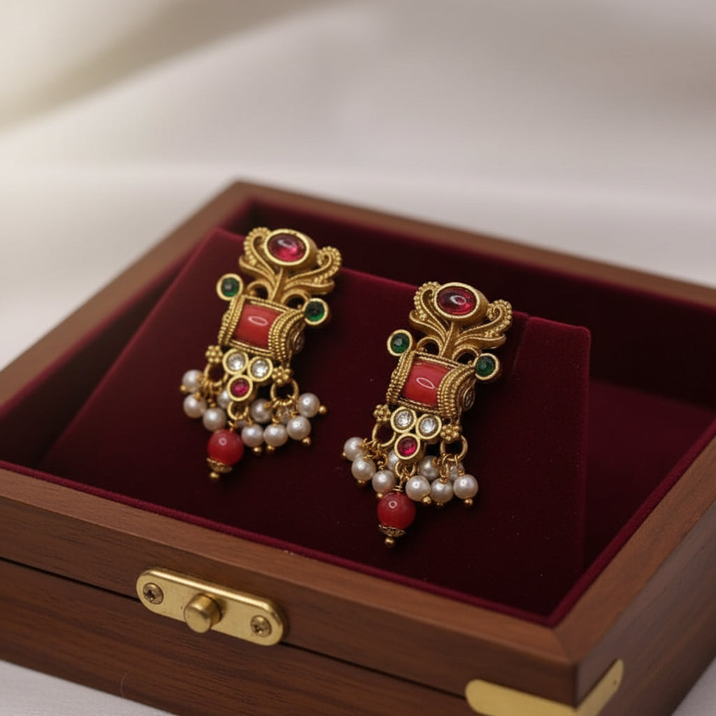 Ornate gold earrings with red and green gemstones on a red velvet surface inside a wooden box.