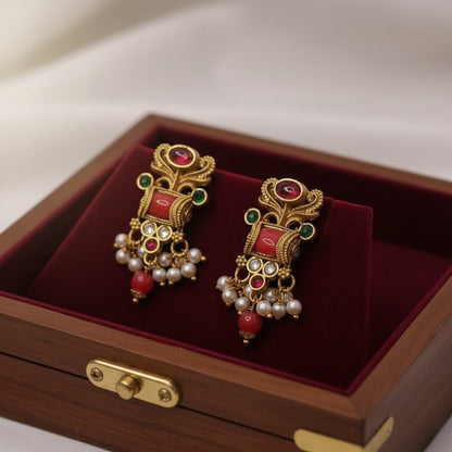 Ornate gold earrings with red and green gemstones on a red velvet surface inside a wooden box.