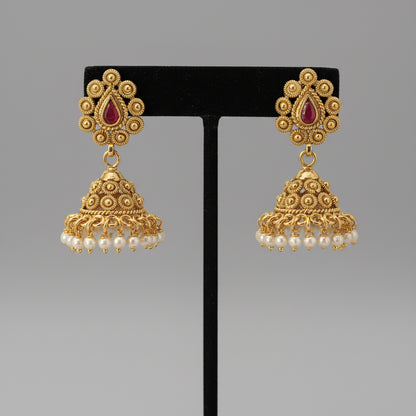 Pair of gold earrings with red stones and pearl drops on a black stand against a gray background