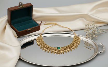 Gold necklace with a green stone on a silver tray with a wooden jewelry box and white flowers.