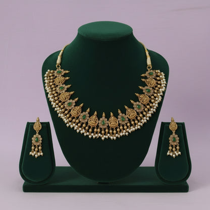 Gold necklace with green stones and matching earrings on a green stand against a light purple background