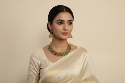 Woman wearing a white saree with gold border and green jewelry on a beige background