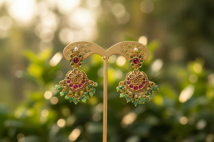 Gold earrings with green and red stones on a gold stand against a beige background