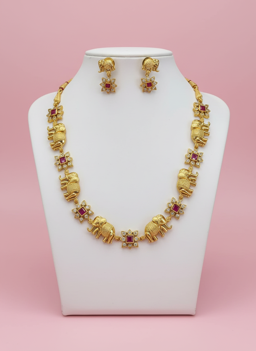 Gold necklace with elephant and ruby designs on a white stand against a pink background