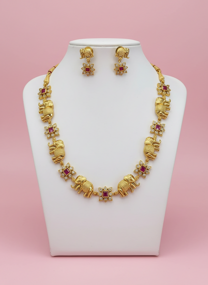 Gold necklace with elephant and ruby designs on a white stand against a pink background