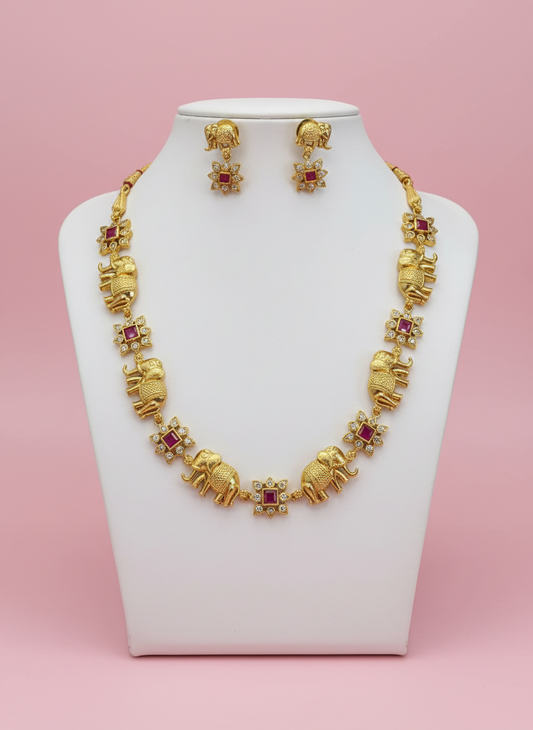 Gold necklace with elephant and ruby designs on a white stand against a pink background