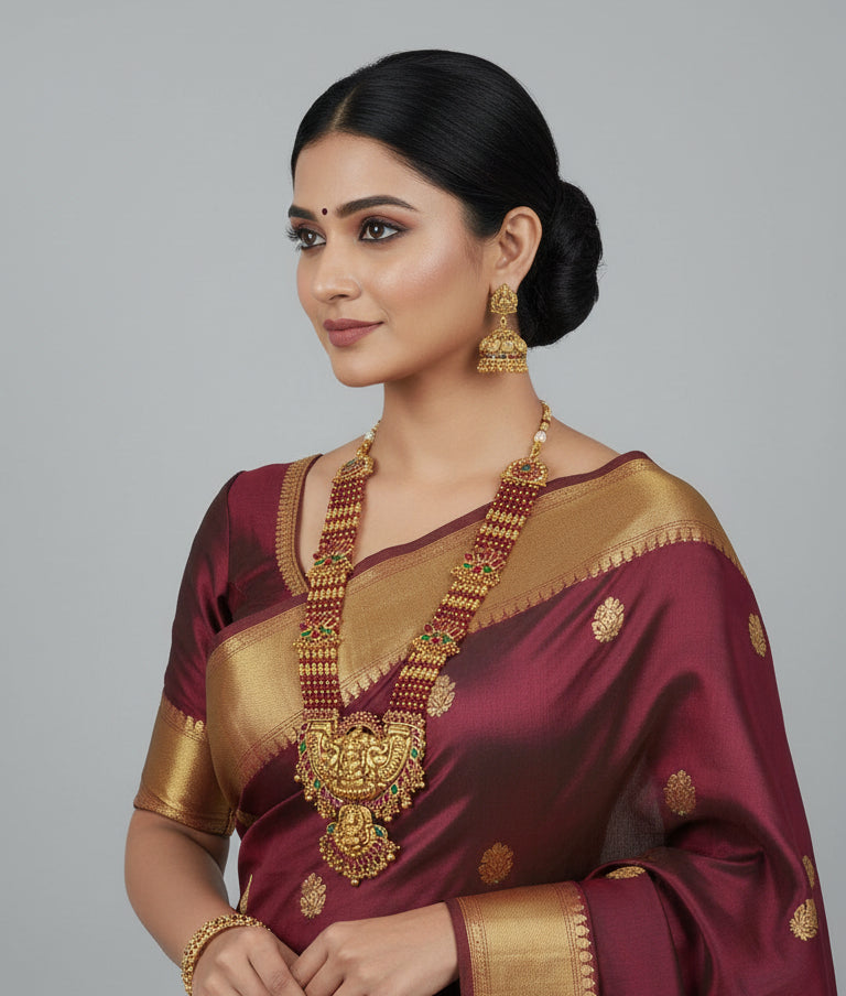 Woman wearing a traditional maroon saree with gold border and jewelry on a gray background