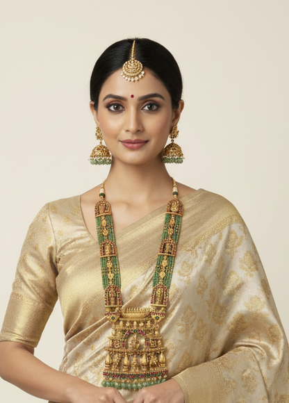 Woman wearing traditional gold and green jewelry with a beige saree against a plain background