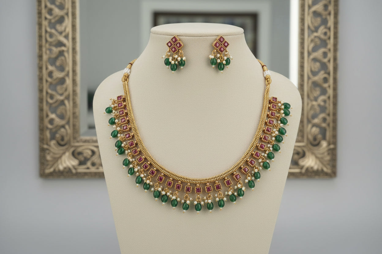 Gold necklace with green and red beads on a mannequin bust, with matching earrings in the background.