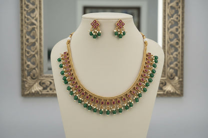 Gold necklace with green and red beads on a mannequin bust, with matching earrings in the background.