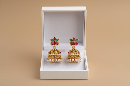 Gold earrings with red and green accents in a white box on a beige background