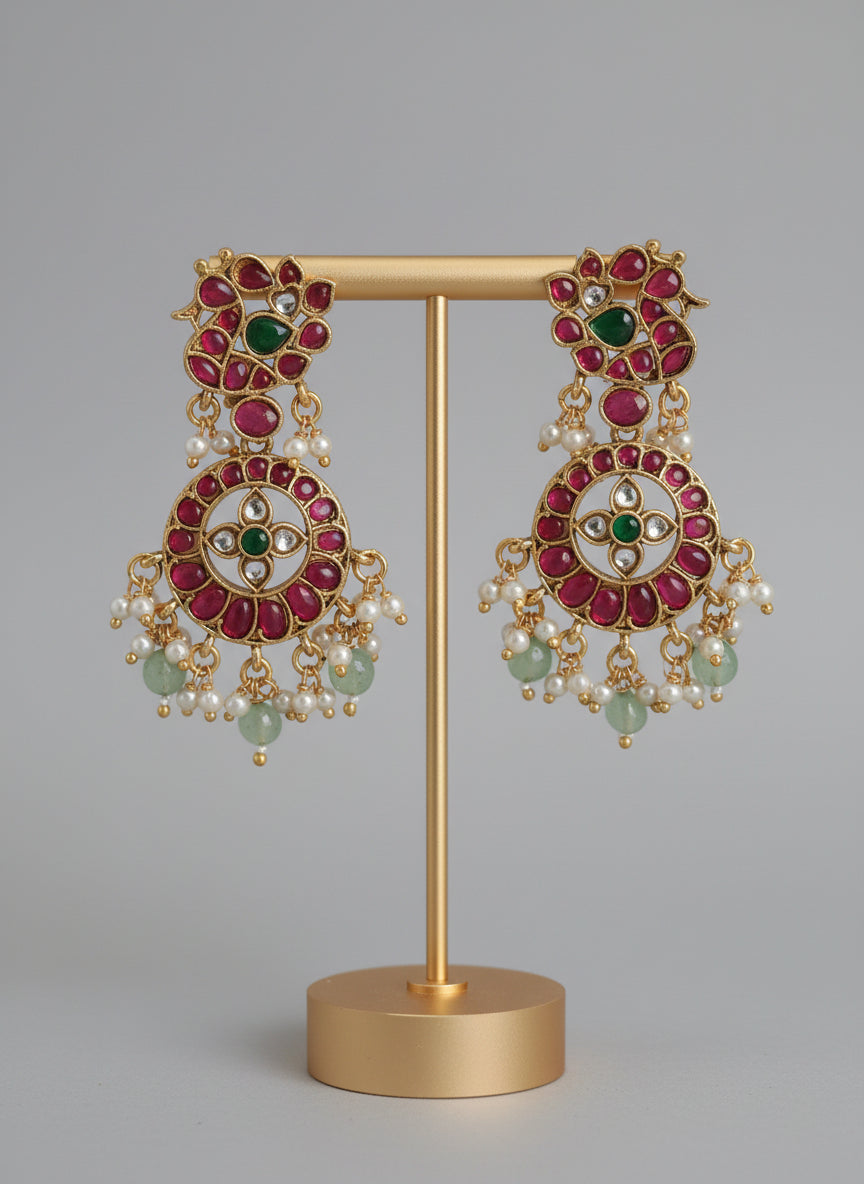 Ornate earrings with red, green, and pearl accents on a gold stand against a gray background