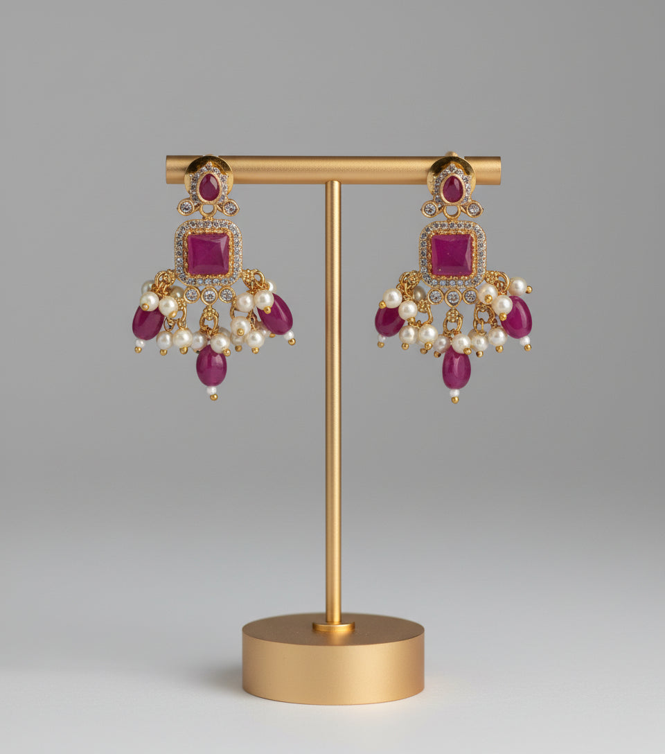 Pair of gold earrings with red stones and pearls on a gold stand against a gray background