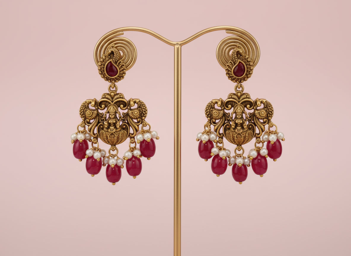 Ornate gold earrings with red gemstones and pearl-like beads on a pink background