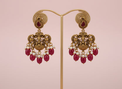 Ornate gold earrings with red gemstones and pearl-like beads on a pink background