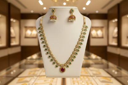Gold necklace with green gemstones and red stones on a white stand against a beige background