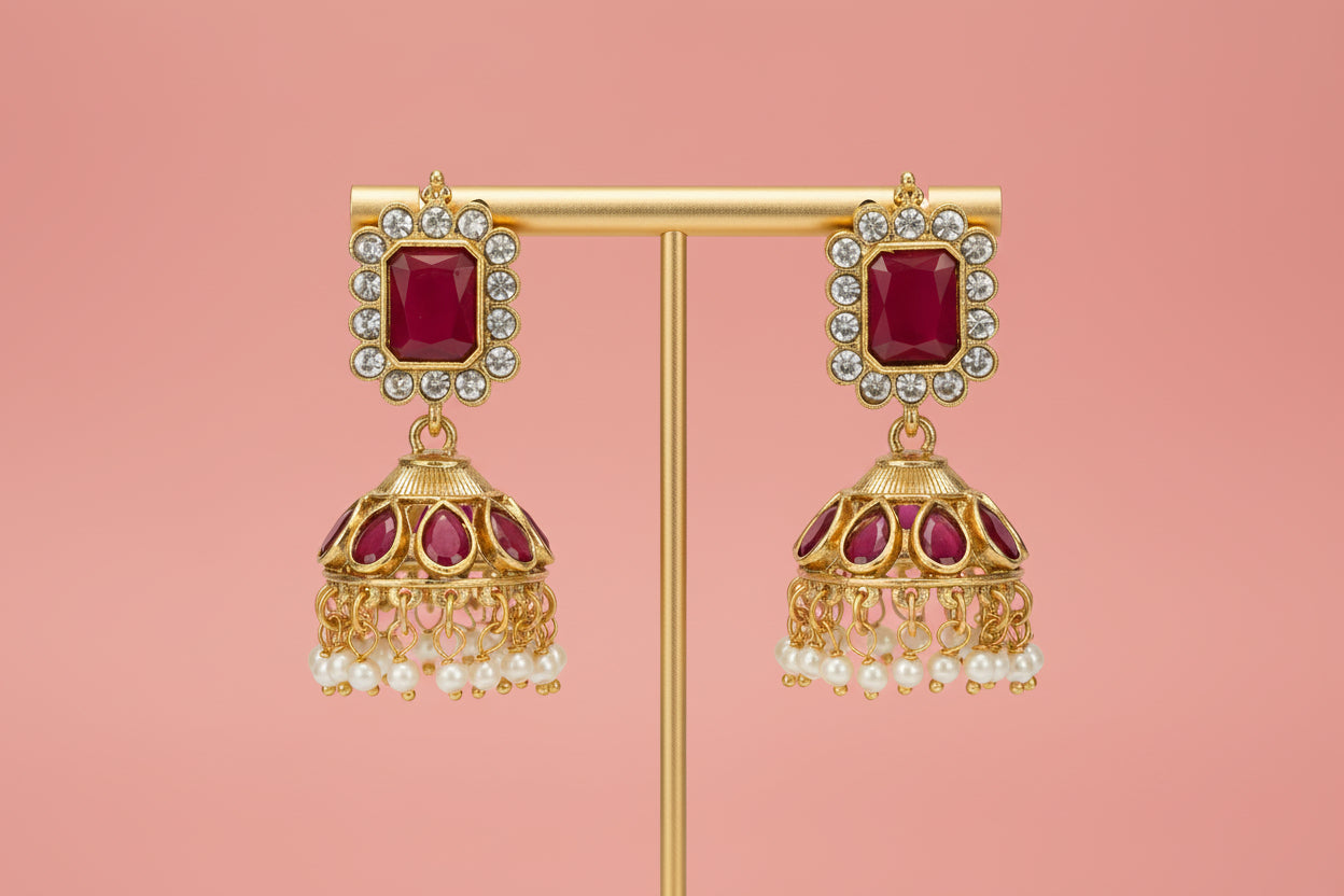 Gold earrings with red gemstones and pearl-like elements on a stand against a light gray background