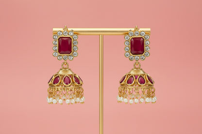 Gold earrings with red gemstones and pearl-like elements on a stand against a light gray background