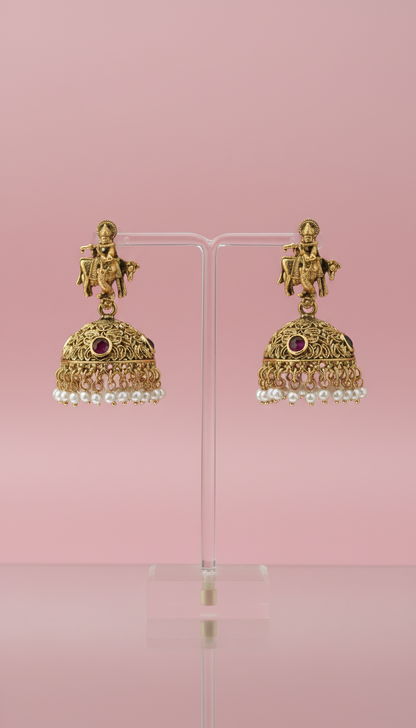 Gold earrings with pearls and red stones on a clear stand against a pink background