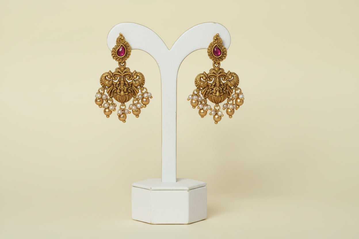Ornate gold earrings with red gemstones on a white stand against a pink background